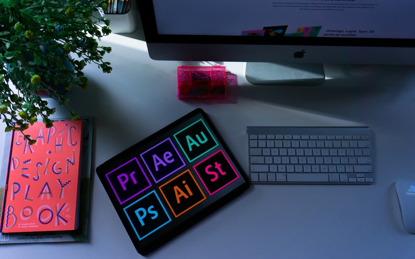 Mesa de trabajo con una tableta mostrando iconos de programas de diseño, un libro de diseño gráfico y una planta